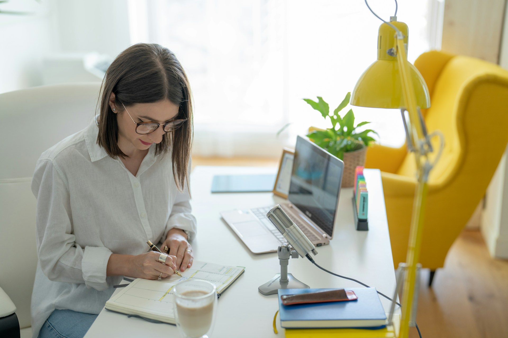 Una mujer con gafas se sienta en un escritorio escribiendo en un cuaderno. Está rodeada de una computadora portátil, un teléfono inteligente, una lámpara amarilla, una maceta, un micrófono y una taza de café, todas herramientas esenciales para crear contenido de calidad.