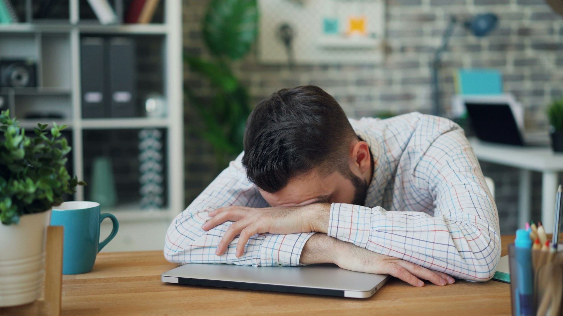 Un hombre con una camisa a cuadros apoya la cabeza sobre los brazos cruzados sobre una computadora portátil cerrada sobre un escritorio, y parece exhausto, tal vez por lidiar con las penalizaciones de Google.