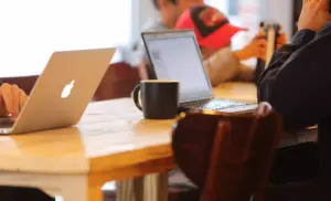 Dos personas trabajan con ordenadores portátiles en una mesa de madera con una taza negra en medio, posiblemente discutiendo sobre arquitectura web para mejorar el SEO; se ven figuras borrosas al fondo.