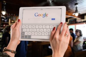 Una persona sostiene una tableta blanca que muestra la página de inicio de la búsqueda de Google, con una escena borrosa de una cafetería interior de fondo.