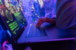 Persona tecleando en un ordenador portátil que muestra un código verde en la pantalla, con un fondo borroso y colorido iluminado por luces azules y moradas.