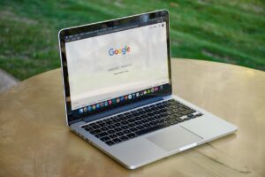 Un ordenador portátil en una mesa redonda al aire libre muestra la página de inicio de Google en un navegador web, con hierba visible en el fondo.