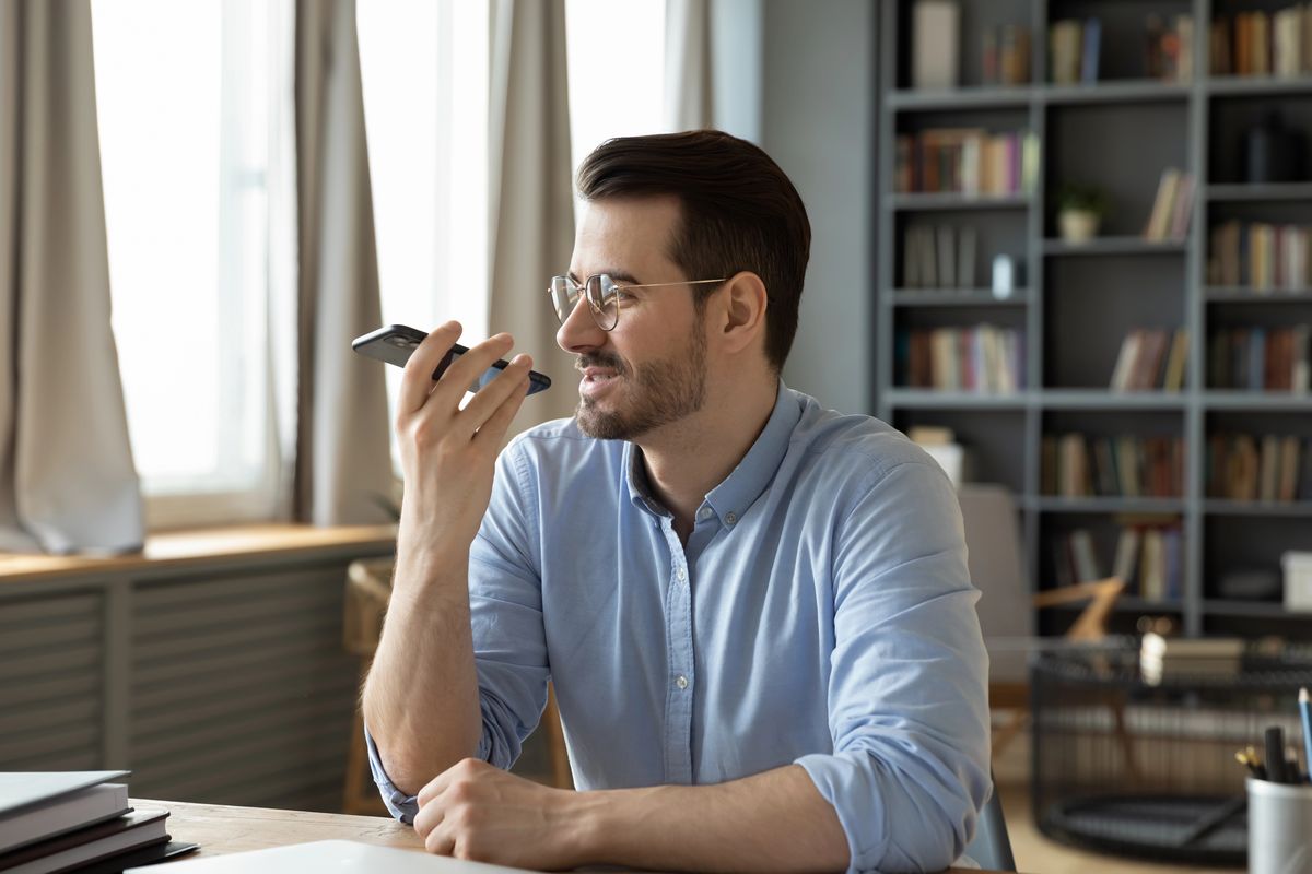 Un hombre con gafas y camisa azul claro está sentado en la mesa de un despacho, con un smartphone en la mano y hablando por él. Al fondo se ven estanterías y grandes ventanales.