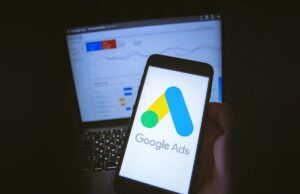 Una mano sostiene un smartphone con el logotipo de Google Ads frente a la pantalla de un portátil que muestra gráficos de análisis.