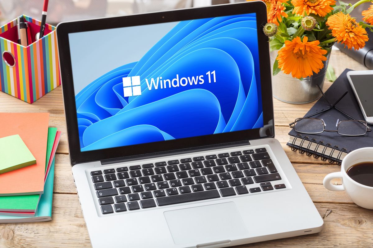 Un portátil con la pantalla de inicio de Windows 11 sobre un escritorio con flores, cuadernos, un smartphone y una taza de café cerca.
