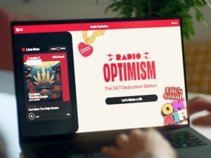 Una persona utiliza un ordenador portátil que muestra un sitio de transmisión de música llamado "Radio Optimism", que muestra una emisora en directo y la opción de hacer una dedicatoria de una canción.