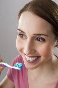 Una mujer sonríe mientras sostiene un cepillo de dientes con pasta dentífrica azul cerca de la boca, preparándose para cepillarse los dientes.