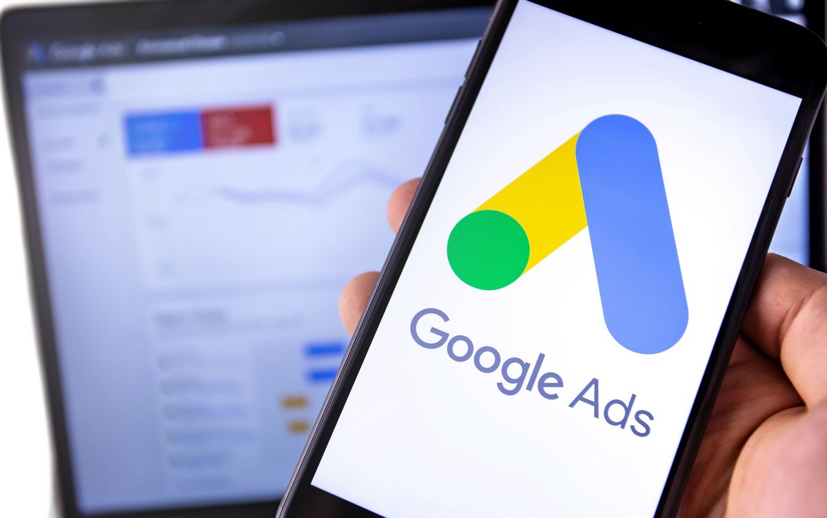 Noticias 1 Una persona sostiene un smartphone en el que aparece el logotipo de Google Ads, con una pantalla de ordenador borrosa que muestra datos analíticos en segundo plano.