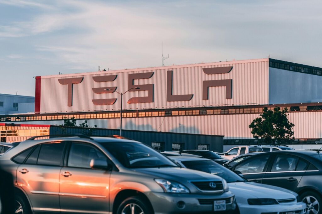 Gran edificio de la fábrica de Tesla con el logotipo destacado al fondo, coches aparcados llenan el primer plano bajo un cielo despejado.