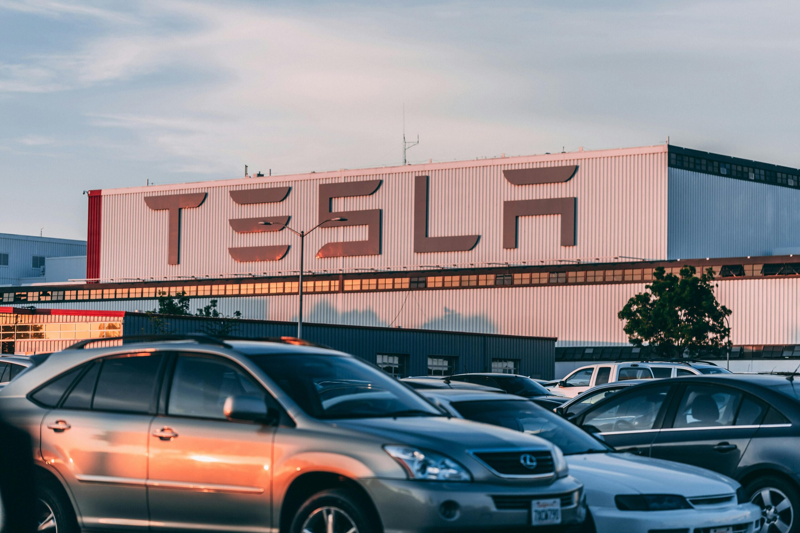 Gran edificio de la fábrica de Tesla con el logotipo destacado al fondo, coches aparcados llenan el primer plano bajo un cielo despejado.