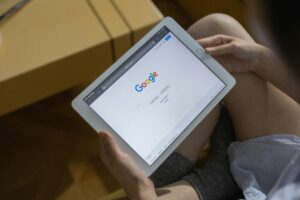 Una persona sostiene una tableta blanca que muestra la página de inicio de la búsqueda de Google, con las piernas visibles en el fondo.