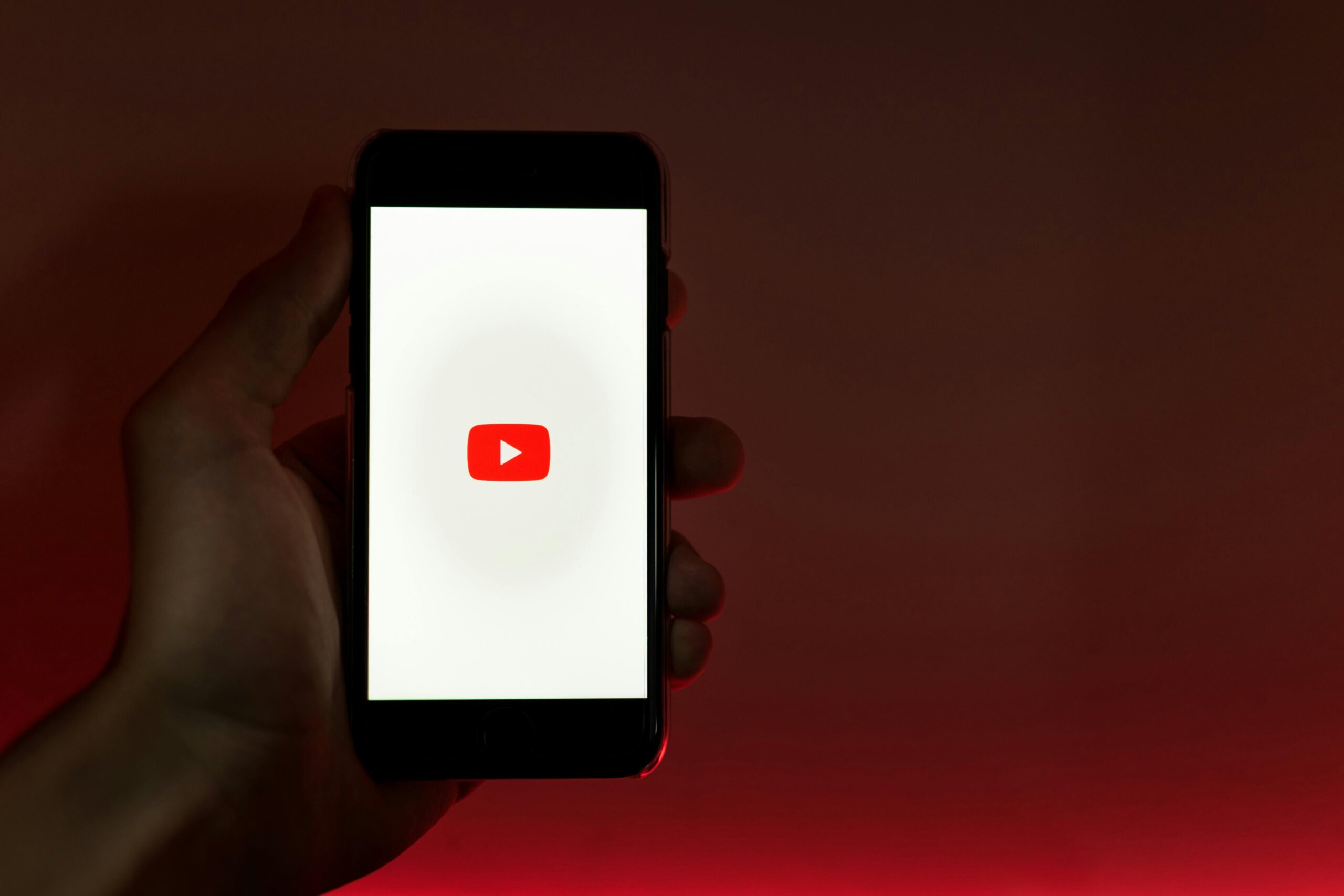 Una mano sostiene un smartphone en cuya pantalla aparece el logotipo de YouTube sobre un fondo degradado en rojo y blanco.