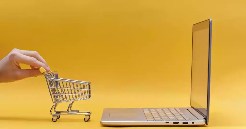 Una mano empuja un pequeño carrito de la compra hacia un portátil abierto sobre un fondo amarillo, simbolizando las compras online y cómo el SEO para eCommerce puede optimizar tu tienda en línea.