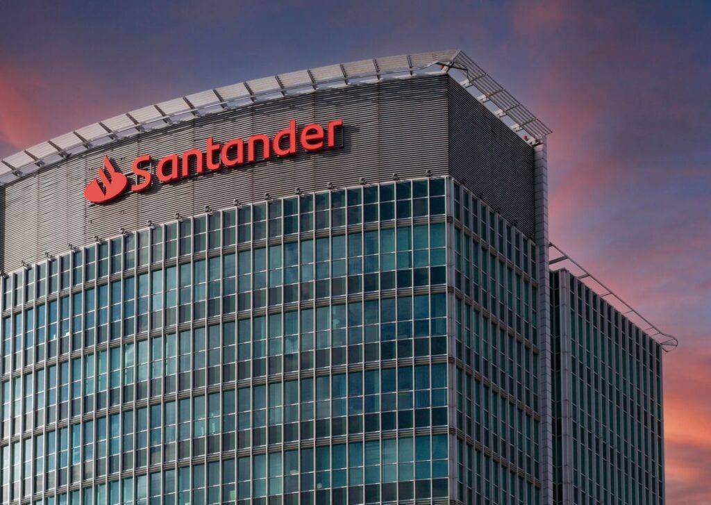 Inicio 8 Un moderno edificio de oficinas de cristal con el logotipo y el nombre del Santander en rojo en la parte superior, fotografiado al atardecer con un cielo rosa y azul de fondo.