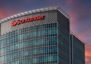 Un moderno edificio de oficinas de cristal con el logotipo y el nombre del Santander en rojo en la parte superior, fotografiado al atardecer con un cielo rosa y azul de fondo.