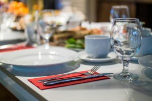Primer plano de una mesa de comedor con un plato blanco, una taza, un tenedor, un cuchillo y una copa de vino sobre una servilleta roja. Otros servicios de mesa y vajilla aparecen difuminados en el fondo.