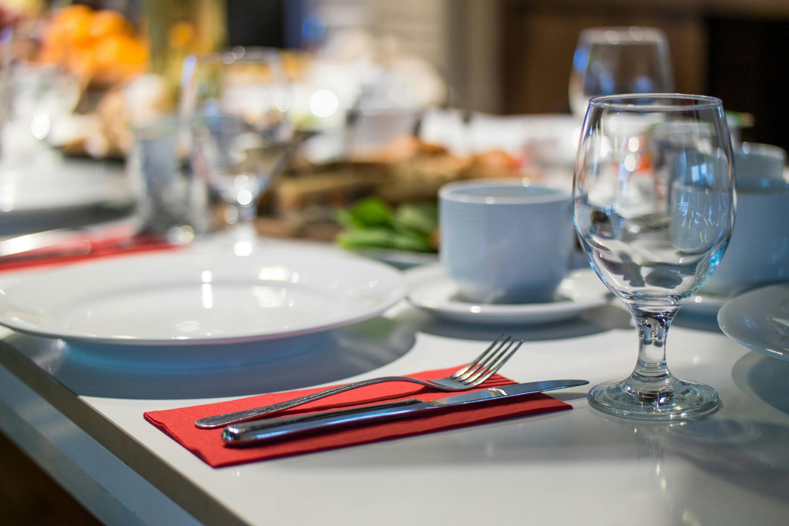 Primer plano de una mesa de comedor con un plato blanco, una taza, un tenedor, un cuchillo y una copa de vino sobre una servilleta roja. Otros servicios de mesa y vajilla aparecen difuminados en el fondo.