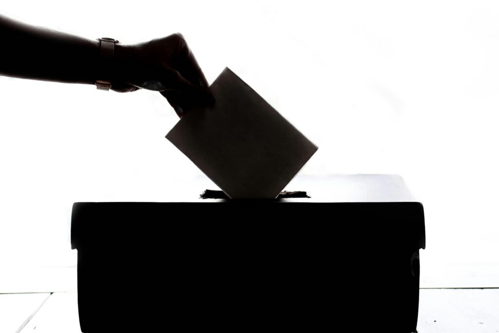 Una mano introduce una papeleta en una urna, silueteada sobre un fondo blanco, representando el acto de votar o la participación electoral.