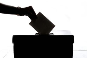 Una mano introduce una papeleta en una urna, silueteada sobre un fondo blanco, representando el acto de votar o la participación electoral.