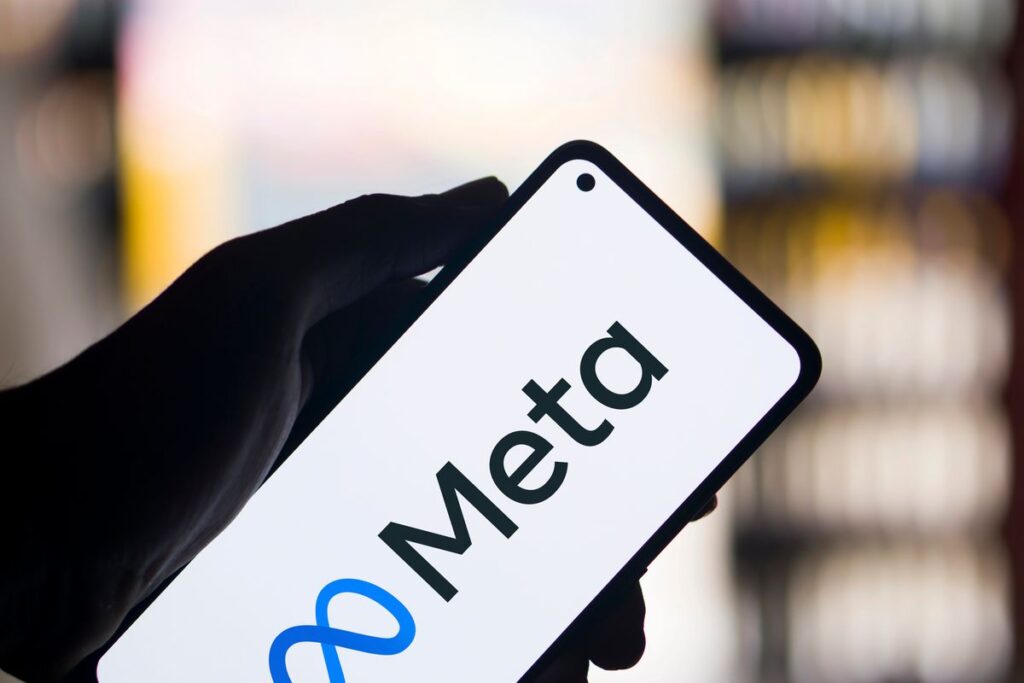 Inicio 8 Una mano sostiene un smartphone que muestra el logotipo y el nombre de Meta en su pantalla, con un fondo borroso de luces.
