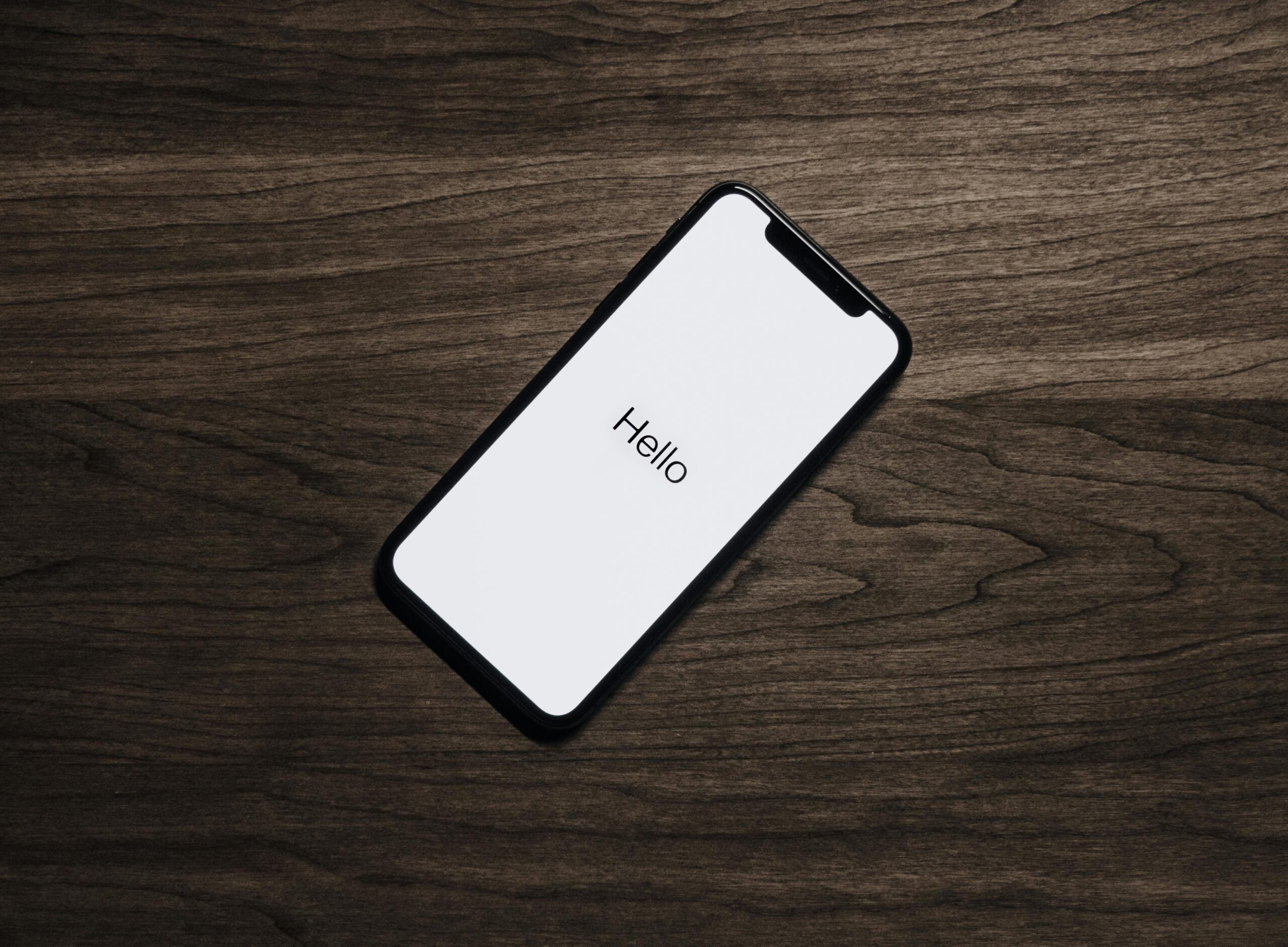 Un smartphone con una pantalla blanca que muestra la palabra "Hola" descansa sobre una superficie de madera oscura.