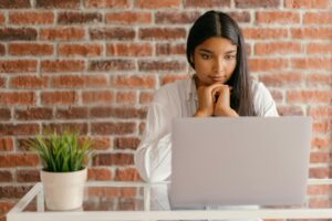 Una mujer está sentada ante un escritorio de cristal, mirando la pantalla de un ordenador portátil con las manos bajo la barbilla. Sobre el escritorio hay una maceta y, al fondo, una pared de ladrillo.