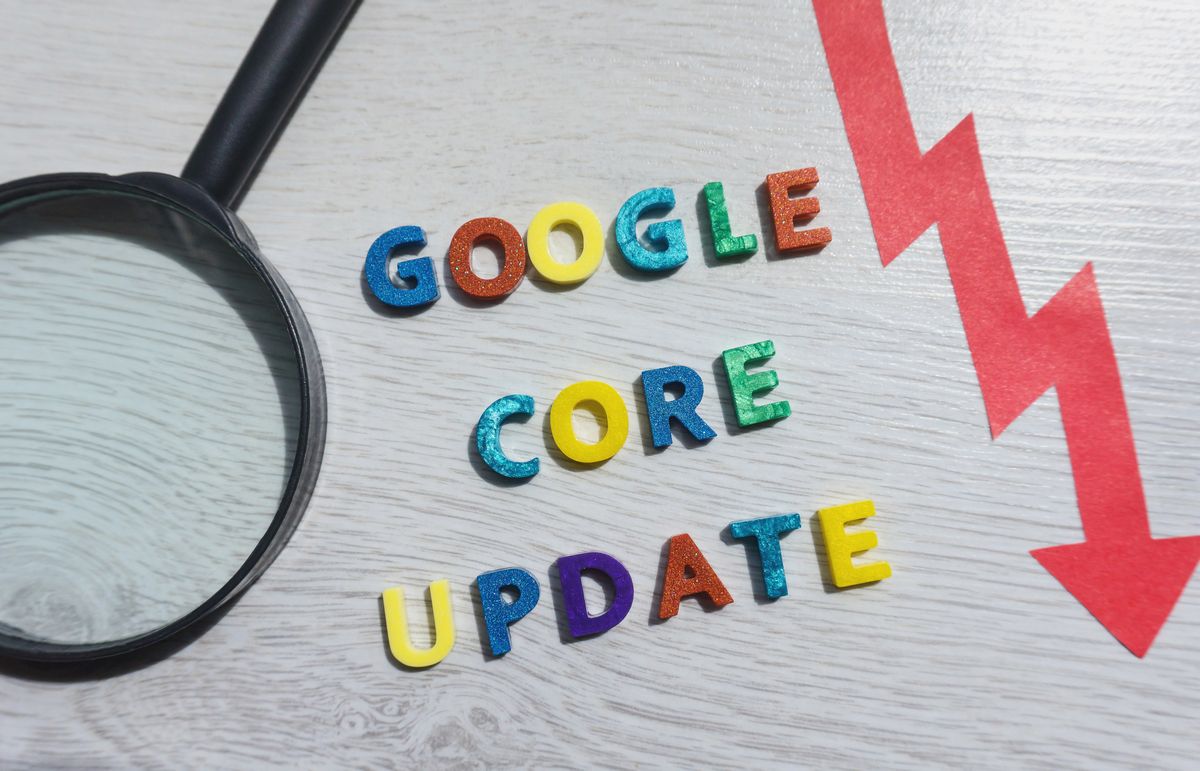 Letras de colores deletrean "Google Core Update" junto a una lupa y una flecha roja hacia abajo sobre una superficie de madera clara.