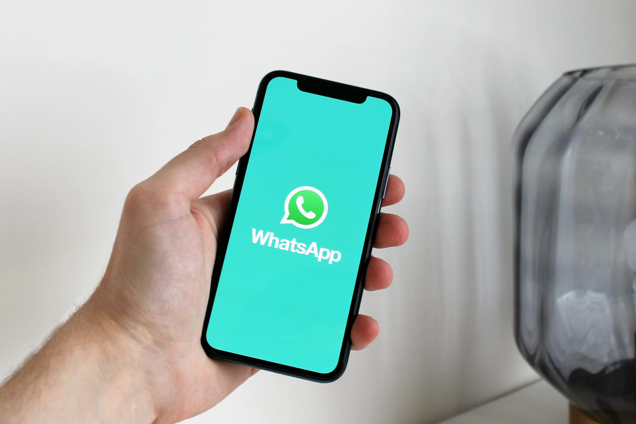 Una mano sostiene un smartphone que muestra el logotipo y el nombre de WhatsApp en su pantalla, con un objeto gris y una pared blanca de fondo.