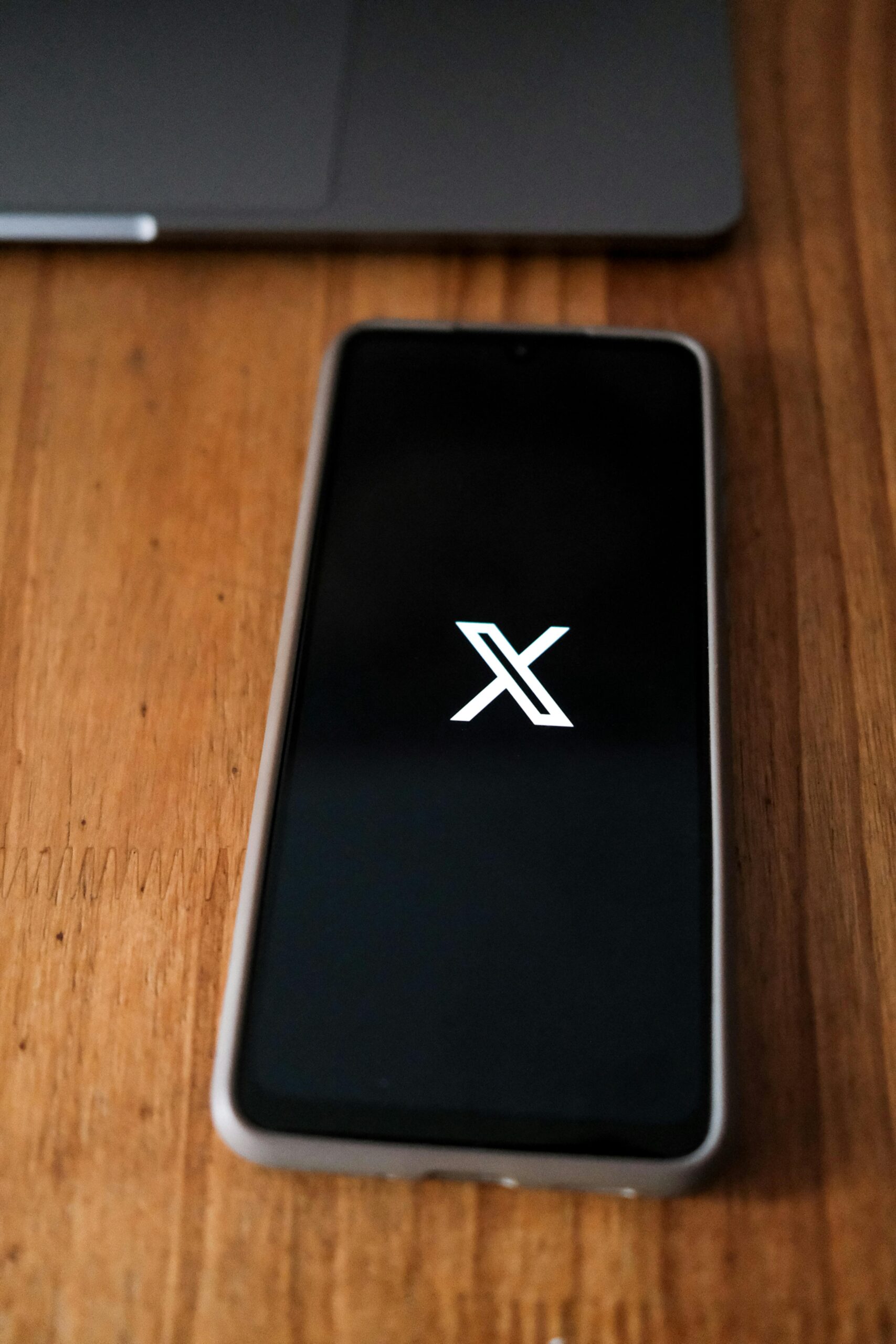 Un smartphone que muestra el logotipo X en su pantalla descansa sobre una superficie de madera, con parte de un ordenador portátil visible en el fondo.