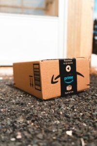 Un paquete de cartón de Amazon Prime se encuentra en una superficie de grava fuera de una puerta, mostrando el logotipo de Prime y la marca.