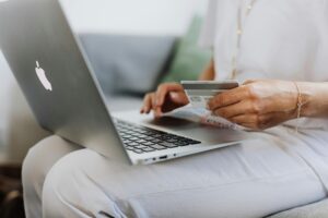 Una persona está sentada con un ordenador portátil en el regazo, sostiene una tarjeta de crédito e introduce información, sugiriendo compras o pagos en línea.