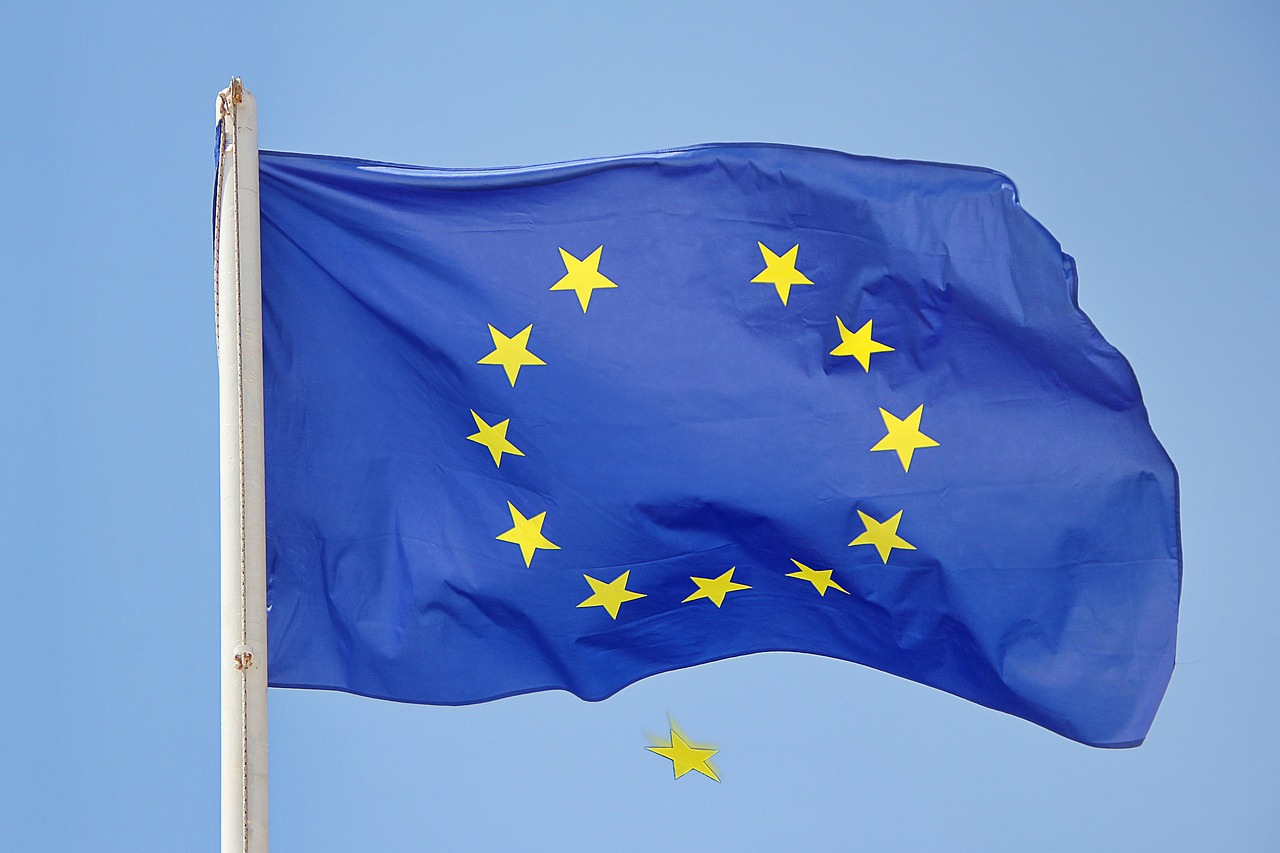 Una bandera de la Unión Europea ondea al viento contra un cielo azul, con una estrella amarilla desprendida y cayendo bajo la bandera.