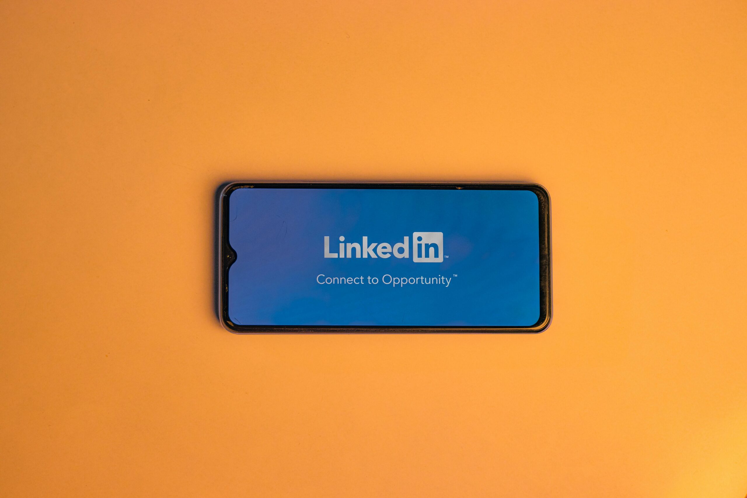 Un smartphone muestra el logotipo de la aplicación LinkedIn y el eslogan "Connect to Opportunity" en una pantalla azul sobre fondo naranja.