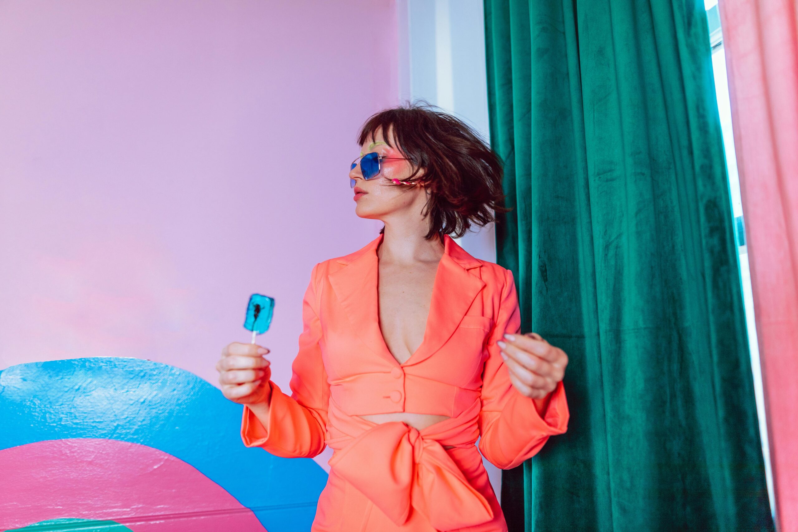 Mujer con traje coral brillante y gafas de sol sostiene un polo, de pie en una colorida habitación con paredes rosas y cortinas verdes.