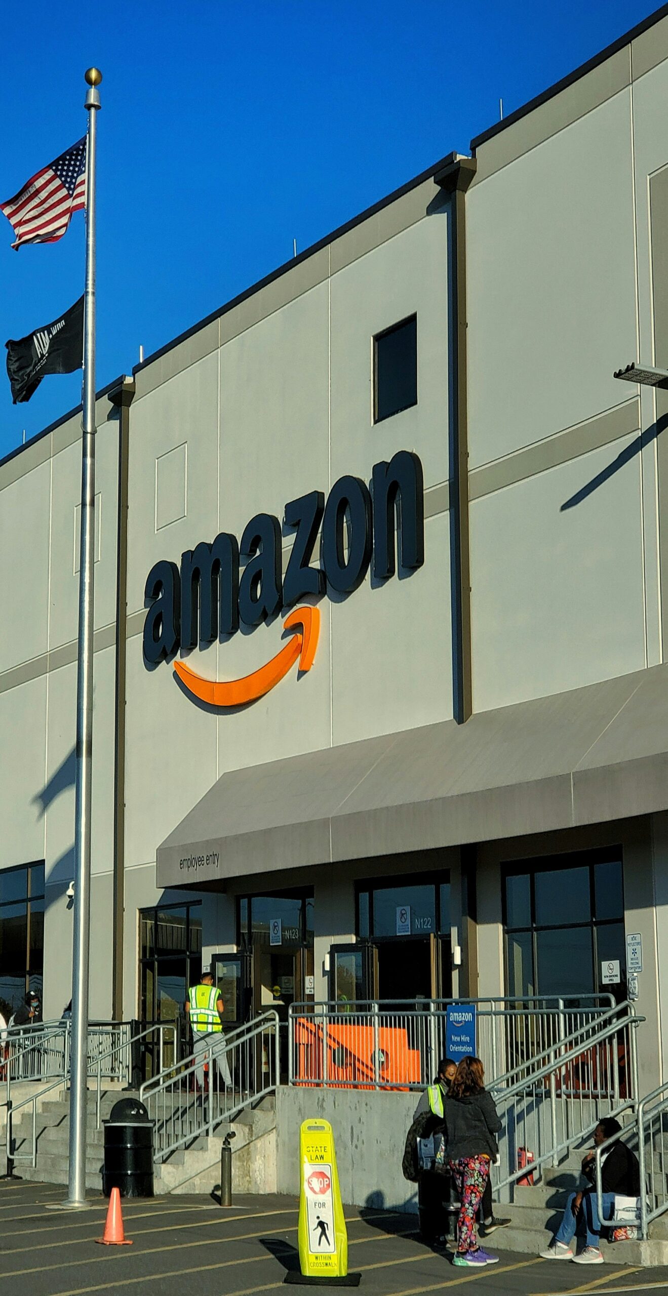 Exterior de un edificio de Amazon con el logotipo de la empresa, una bandera estadounidense y varias personas cerca de la entrada.