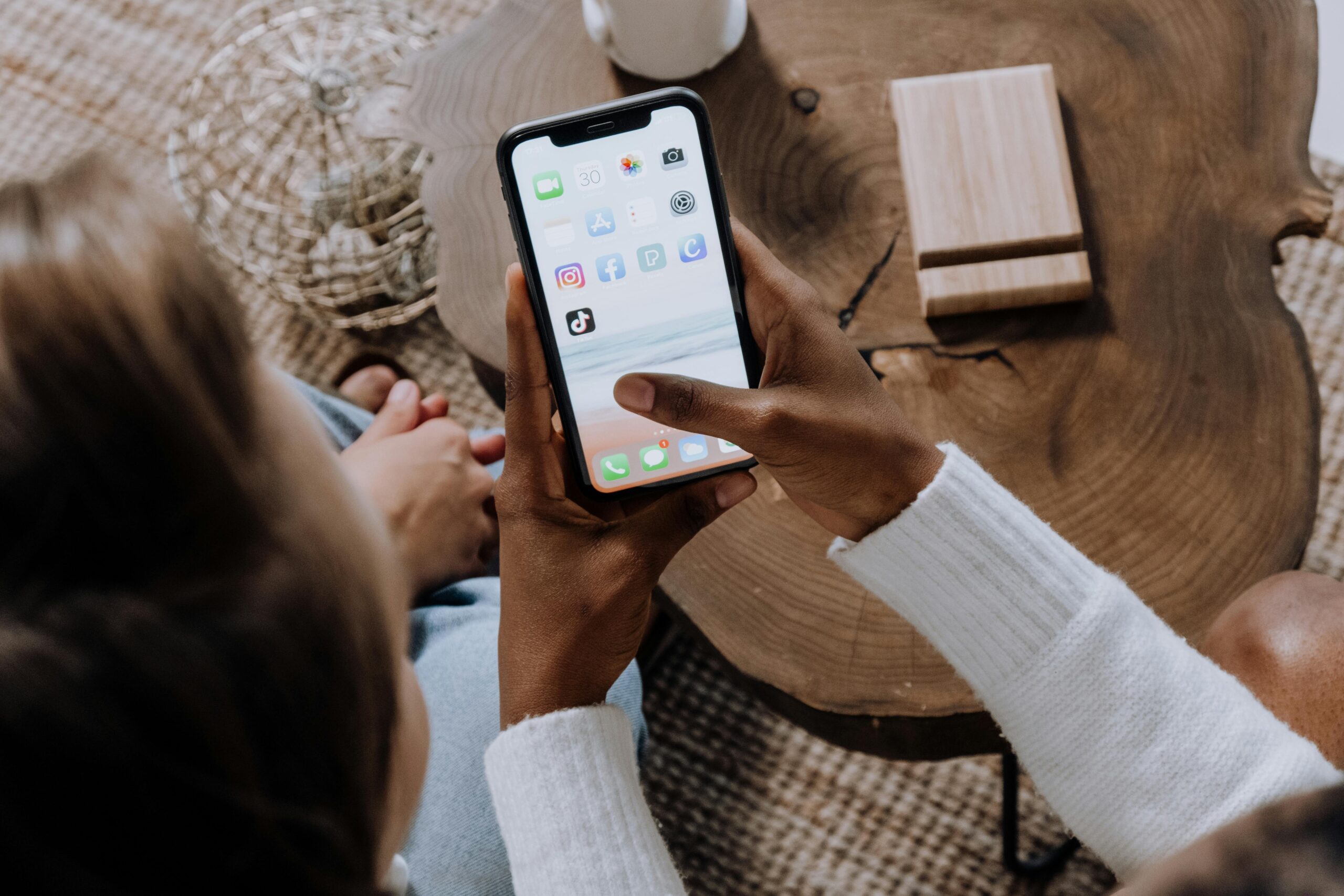 Una persona sujeta un smartphone en el que aparecen varios iconos de aplicaciones de redes sociales, sentada en una mesa de madera con otra persona cerca.