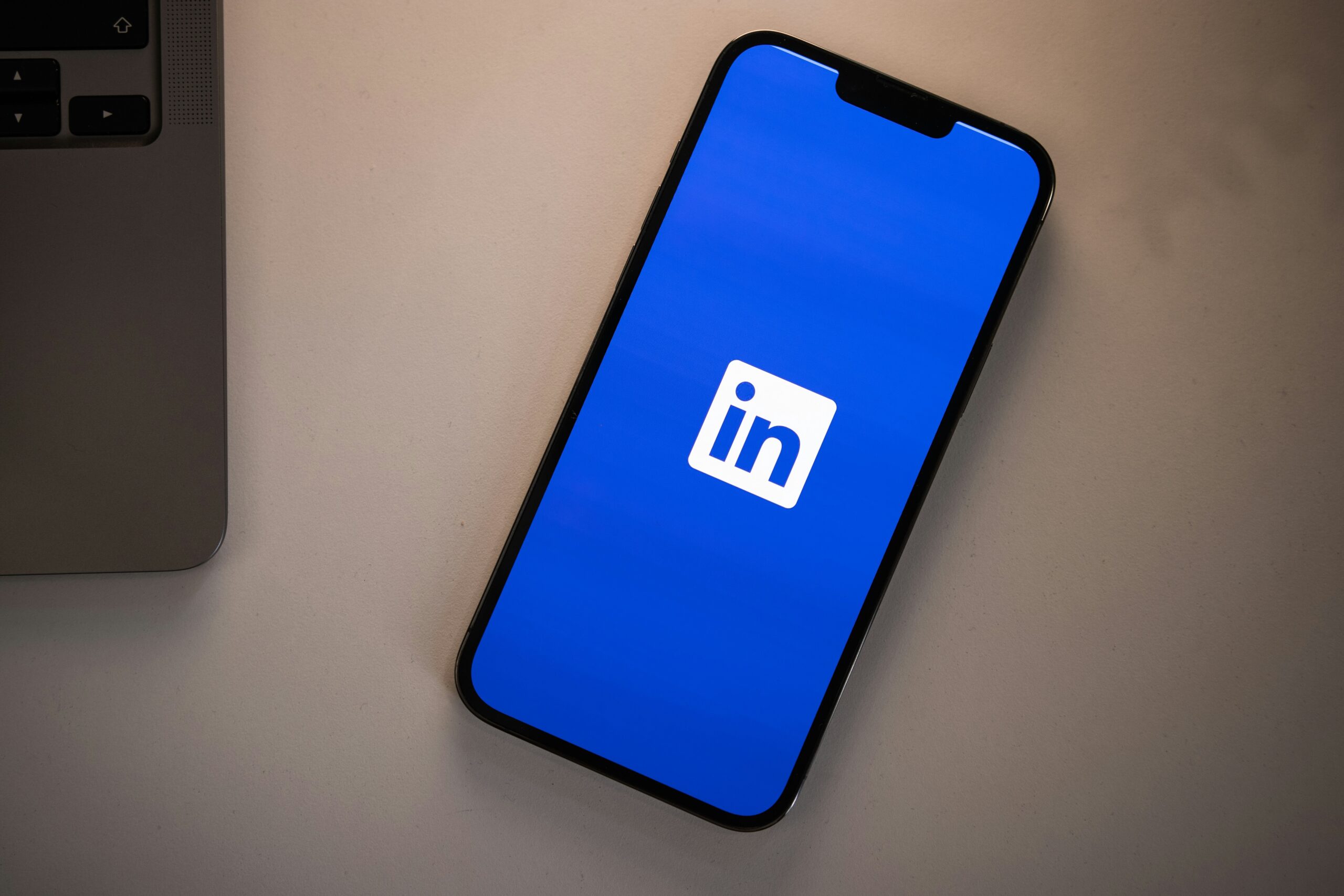 Un smartphone muestra el logotipo de la aplicación LinkedIn en su pantalla, apoyado sobre una superficie blanca junto a la esquina de un ordenador portátil.