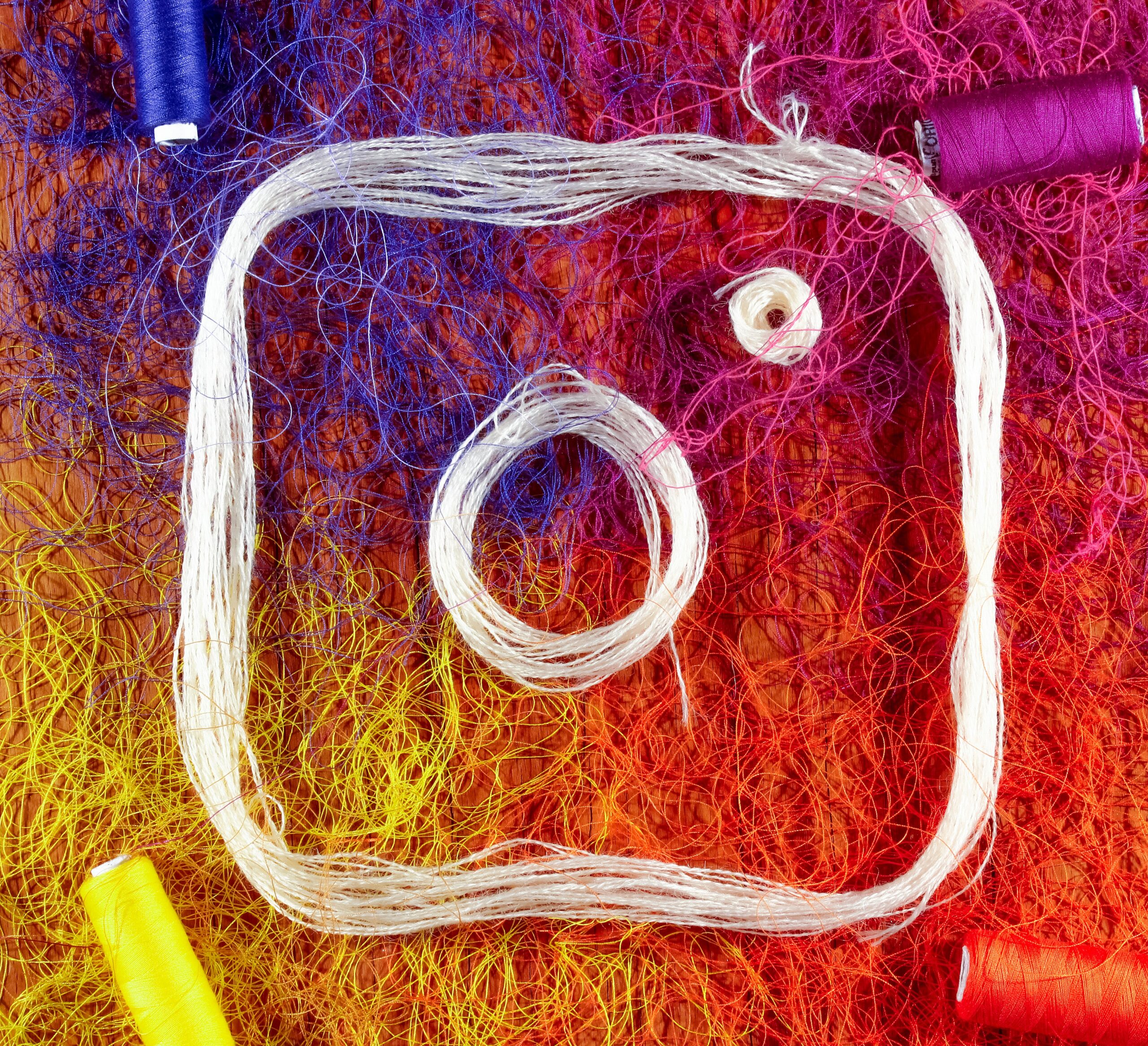 Cuerda blanca dispuesta en forma del logotipo de Instagram sobre un fondo colorido de hilos enredados con carretes de hilo en los bordes.