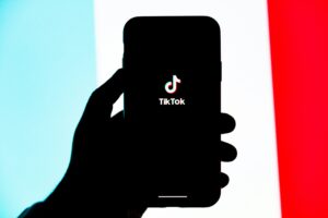 Una mano sostiene un smartphone que muestra el logotipo de TikTok en la pantalla, con un fondo difuminado en azul claro, blanco y rojo.