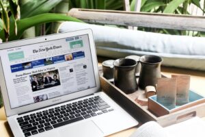 Sobre una mesa, junto a una bandeja con tazas y paquetes de té, un ordenador portátil con la página web de The New York Times, plantas y un cojín al fondo.