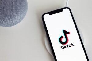 Un smartphone con el logotipo de la aplicación TikTok descansa sobre una plataforma de carga inalámbrica junto a un altavoz inteligente gris sobre una superficie blanca.