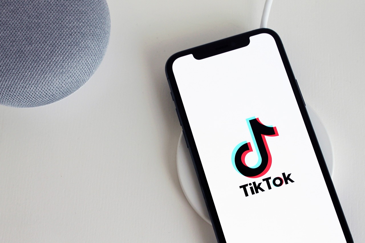 Un smartphone con el logotipo de la aplicación TikTok descansa sobre una plataforma de carga inalámbrica junto a un altavoz inteligente gris sobre una superficie blanca.