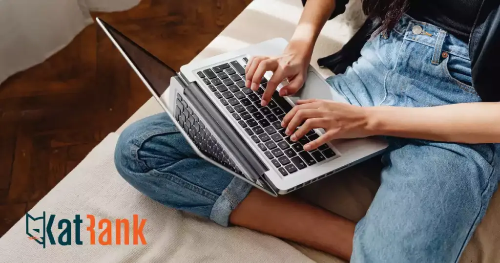 Persona sentada con las piernas cruzadas en un sofá escribiendo en un portátil, con el logotipo de KatRank en la esquina inferior izquierda: perfecto para aprender SEO básico o descubrir cómo hacer una auditoría SEO.
