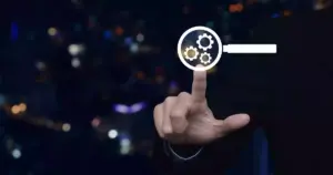 Una persona vestida de traje toca una pantalla virtual en la que aparece una lupa con engranajes, símbolo de la optimización técnica o el análisis impulsado por respuestas de IA, sobre un fondo urbano oscuro y borroso.
