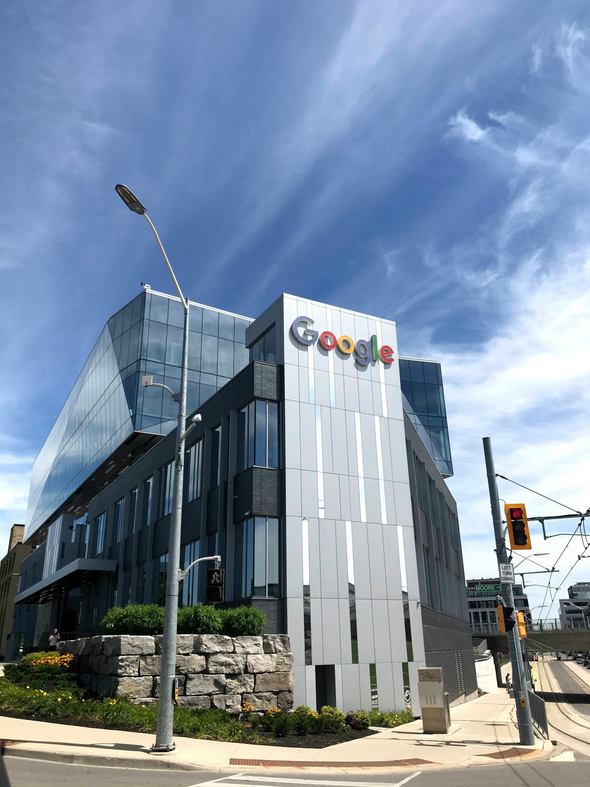 Un moderno edificio de oficinas de Google con ventanas de cristal y un prominente logotipo de Google en la fachada, bajo un cielo parcialmente nublado en un cruce de calles.