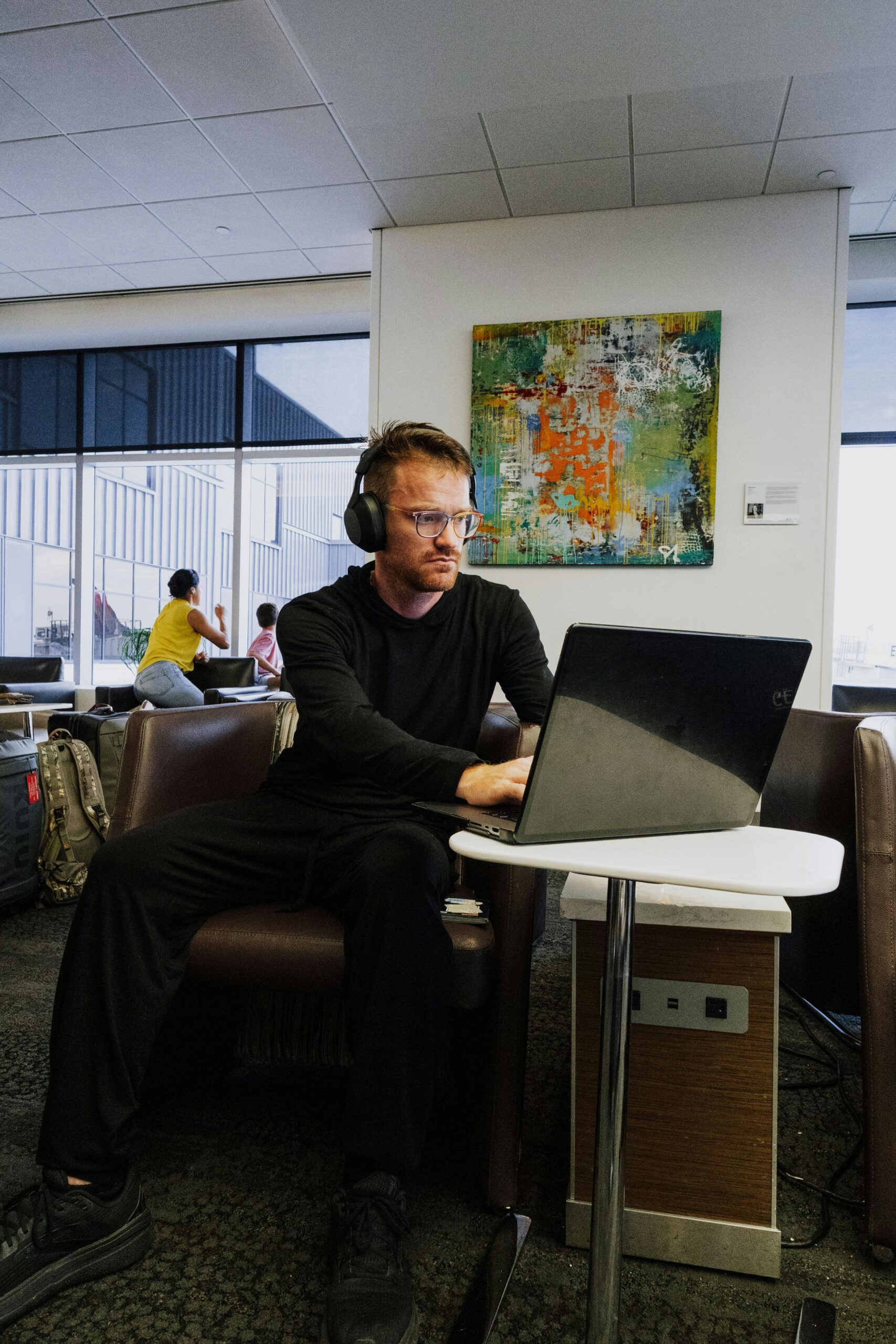 Un hombre con auriculares trabaja con un portátil en la sala VIP de un aeropuerto, con el equipaje y otros viajeros visibles al fondo.