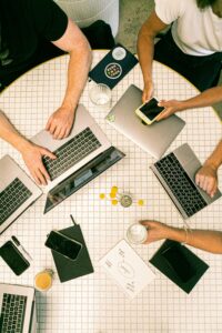 Cuatro personas trabajando alrededor de una mesa redonda con ordenadores portátiles, cuadernos, bebidas y un teléfono, visto desde arriba.
