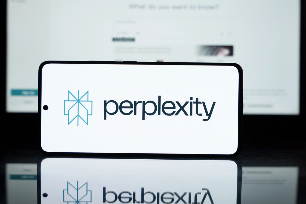 Un smartphone muestra el logotipo y el texto de Perplexity AI, con una pantalla de ordenador borrosa visible en el fondo.