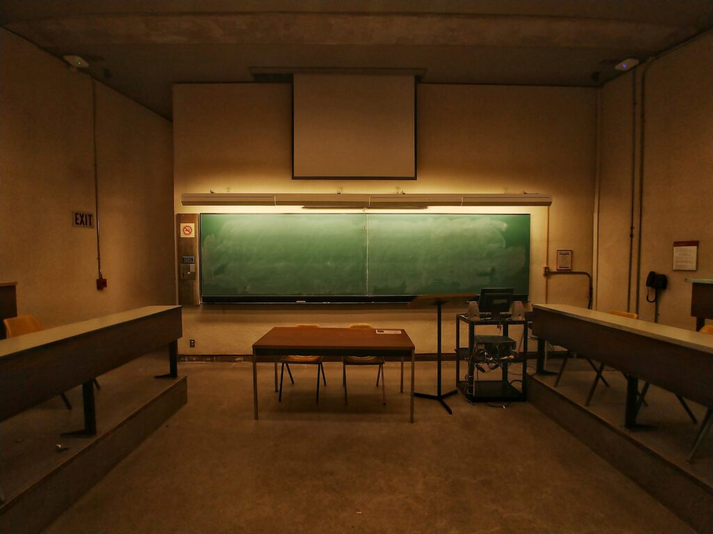 Aula vacía con una gran pizarra verde, escritorio de madera, sillas, podio y equipo informático en la parte delantera.