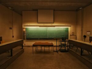 Aula vacía con una gran pizarra verde, escritorio de madera, sillas, podio y equipo informático en la parte delantera.
